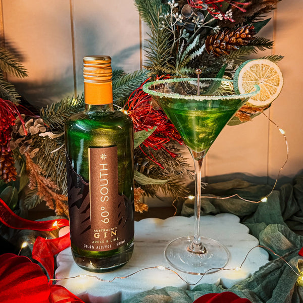 Bottle of 60 South Green Apple and Lime Glitter Gin with a green cocktail garnished with a lime wedge, set against a festive background.