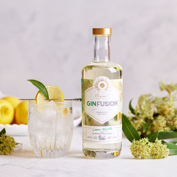 A bottle of GinFusion Lemon Myrtle with Elderflower Tonic next to a glass of the tonic with ice and a lemon slice garnish, surrounded by lemon myrtle leaves and flowers.