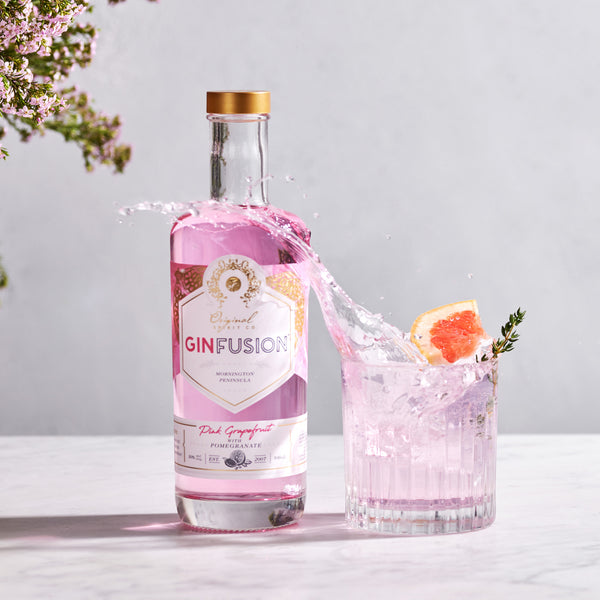 A bottle of Ginfusion Pink Grapefruit with Pomegranate gin, alongside a glass with ice and a slice of grapefruit making a dynamic splash. The background includes a touch of greenery.