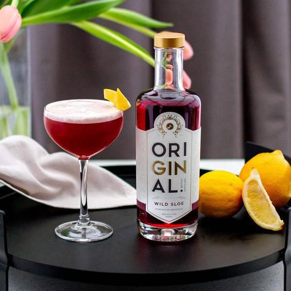 A bottle of Original Wild Sloe Gin with a glass of sloe gin cocktail, lemon slices, and a key ring on a table.