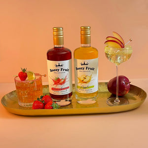 A bottle of Boozy Fruit Strawberry and Apple Vodka on a tray, with cocktails surrounded by fresh strawberries and red apple, showcasing its vibrant colours.