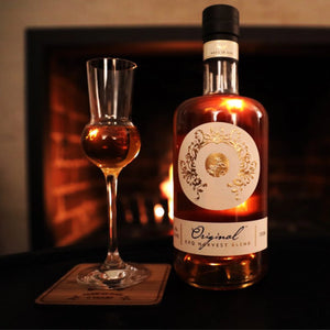 Close-up image of Original Spirit Co.'s EXQ Harvest GinFusion bottle with a glass, set against a warm, cosy fireplace background.